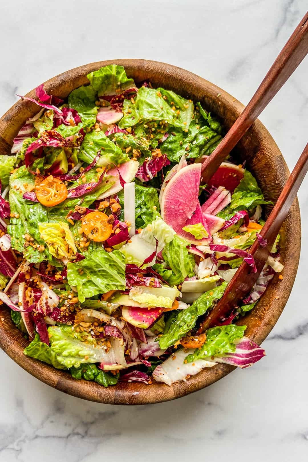 A large wooden salad bowl with a greens, radicchio, kumquats, watermelon radishes, and tongs.