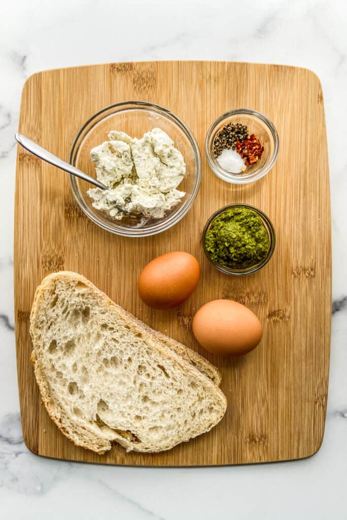 Two slices of sourdough bread, a bowl of goat cheese, two eggs, pesto, and seasonings on a wood cutting board.