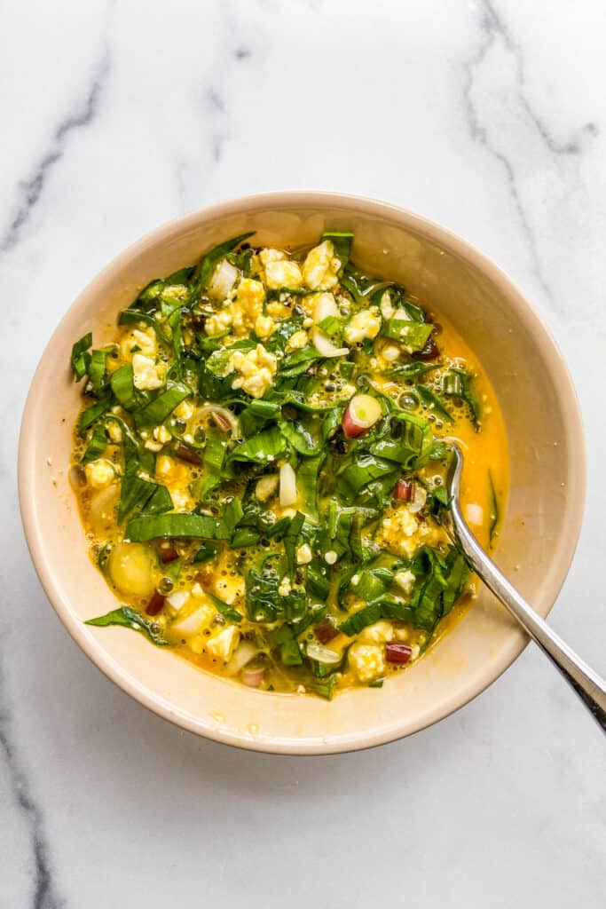 A bowl of beaten eggs with chopped ramps and crumbled feta mixed together.