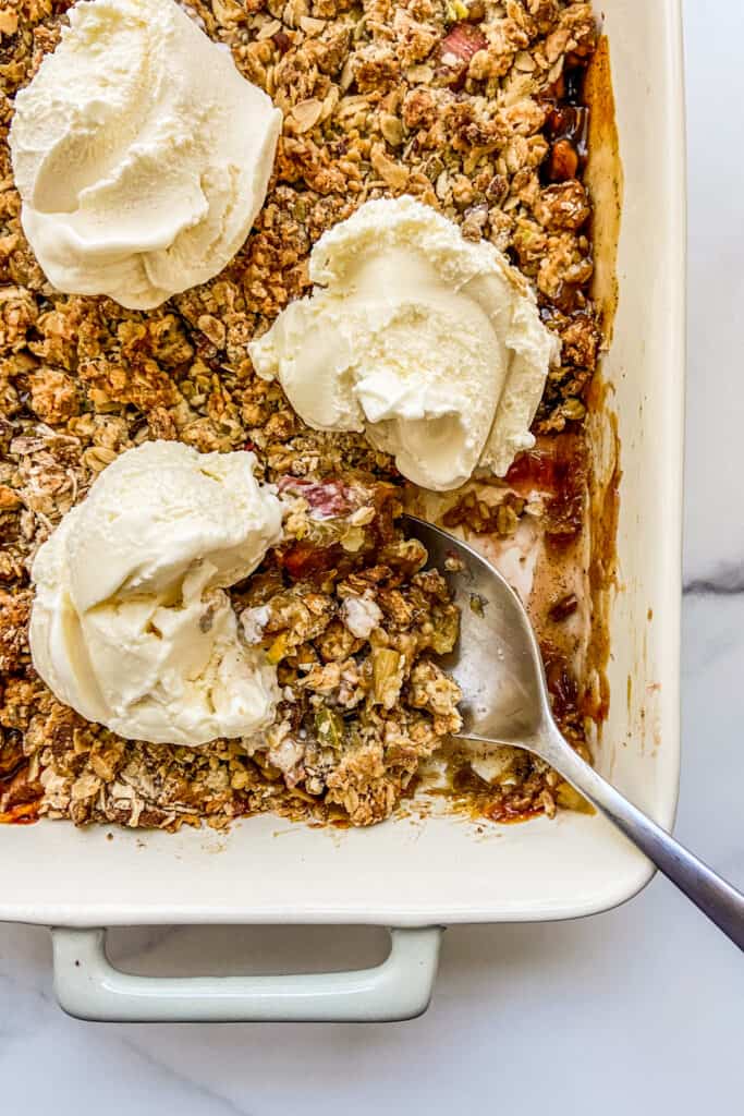 A closeup shot of the edge of a pan of rhubarb crisp, with scoops of vanilla ice cream and a large serving spoon.