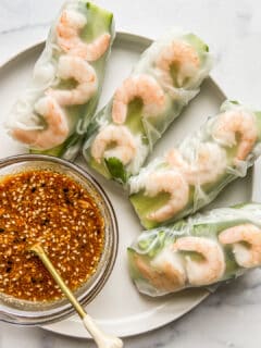 Four shrimp summer rolls on a white plate next to a bowl of soy peanut dipping sauce.