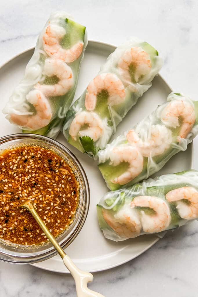 Four shrimp summer rolls on a white plate next to a bowl of soy peanut dipping sauce.