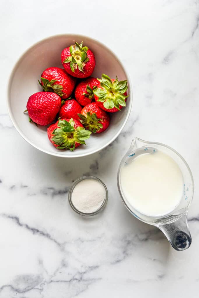 A bowl of strawberries, a measuring cup of milk, and a small bowl of sugar.