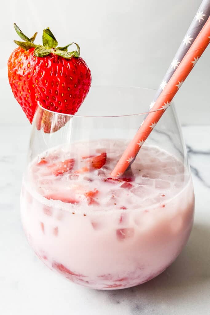 A wine glass with strawberry milk, two straws, and a whole strawberry garnish.