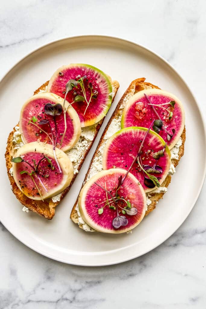 Two slices of toast with goat cheese and watermelon radish on a white plate. 