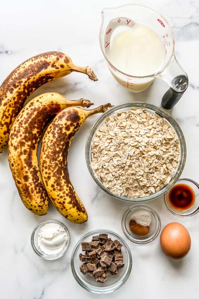 Three bananas, a measuring cup of milk, a bowl of oats, an egg, a bowl of baking powder, a bowl of chocolate chunks, a bowl of cinnamon and salt, and a bowl of vanilla. 