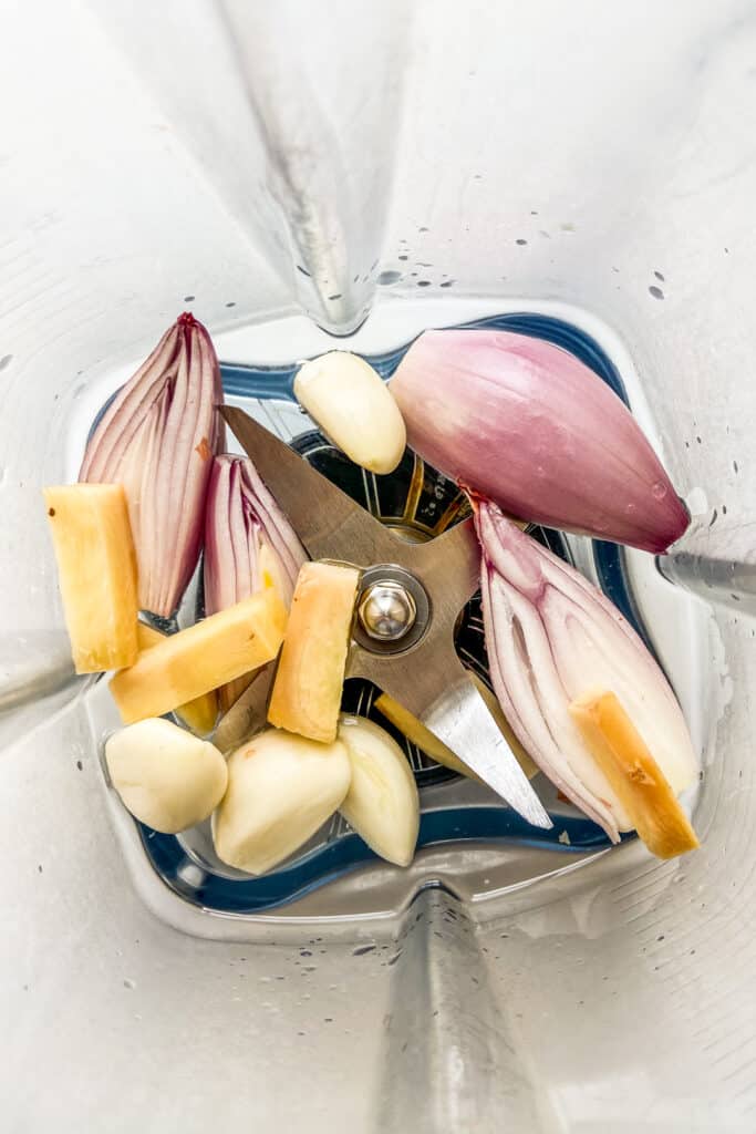 An overhead shot of the inside of a blender with shallots, garlic cloves, and chopped ginger.