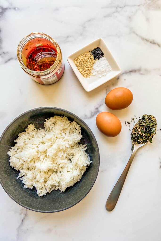 A jar of chili crunch oil, a bowl of sesame seeds, two eggs, a spoon of furikake, and a bowl of rice. 