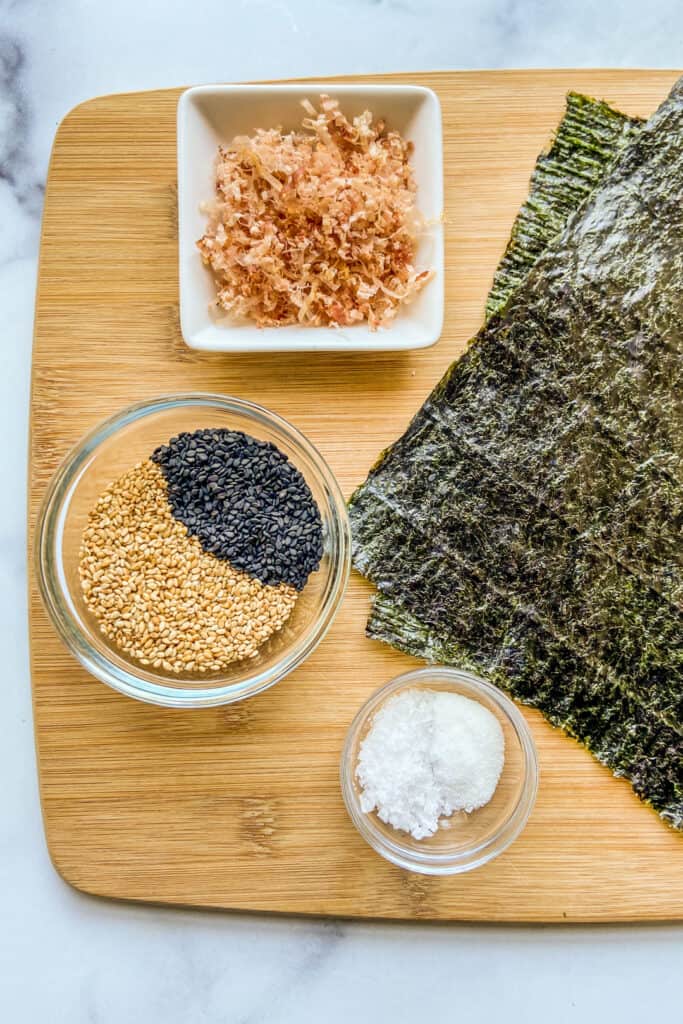 Two nori sheets, a bowl of bonita flakes, a bowl of black and white sesame seeds, and a bowl of salt and sugar on a cutting board.