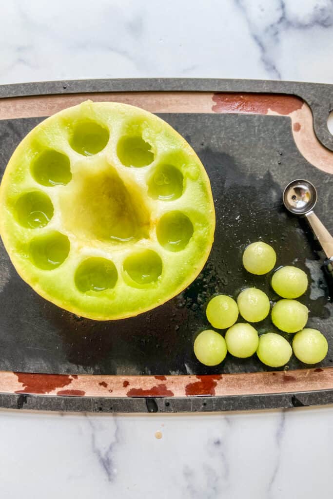 A cutting board with a halved honeydew and melon balls on it.