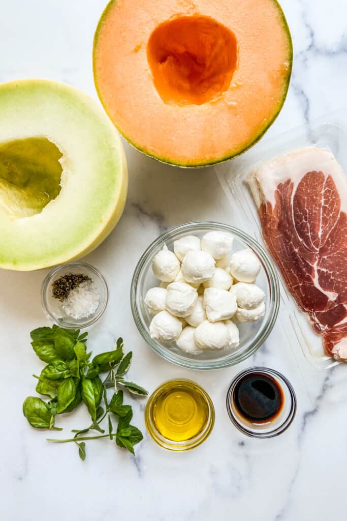 A halved cantaloupe, a halved honeydew, prosciutto, mozzarella balls, fresh herbs, salt, pepper, olive oil, and balsamic.