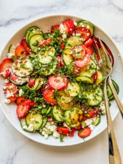 A large white bowl with cucumber strawberry salad topped with herbs and feta.