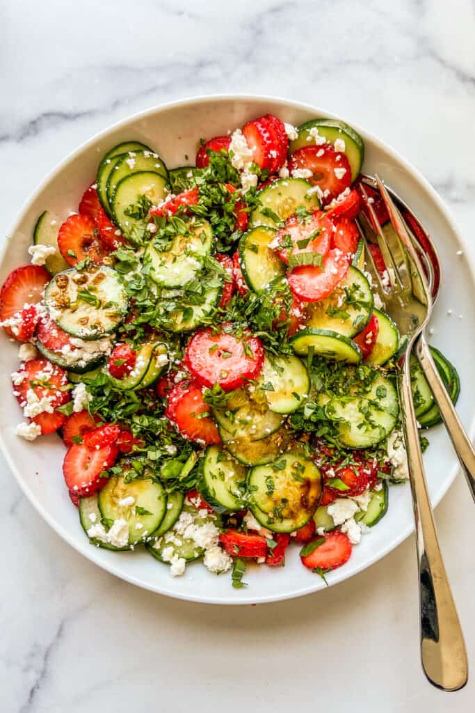 A large white bowl with cucumber strawberry salad topped with herbs and feta.