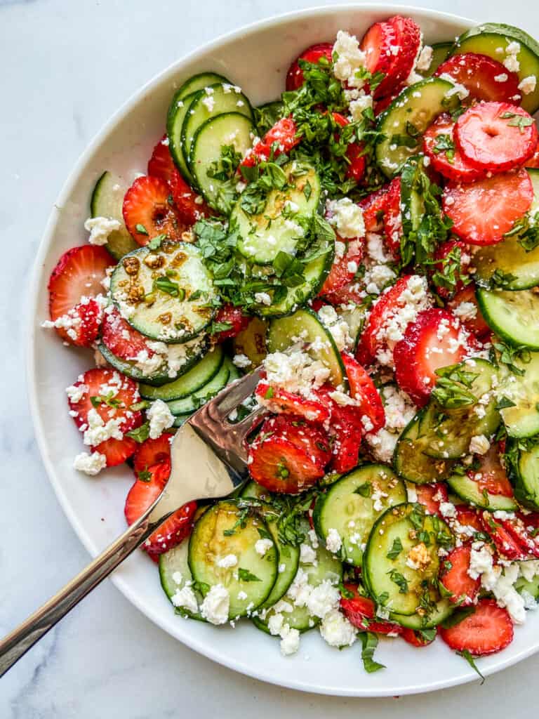 Closeup of a strawberry cucumber salad in a white serving dish with a silver spoon.