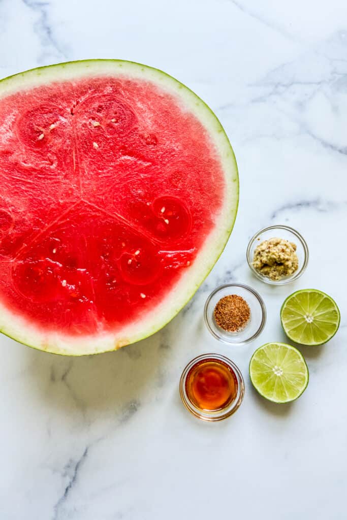 A half a watermelon, a bowl of mustard, a bowl of honey, a bowl of tajin seasoning, and two lime halves on a marble background.