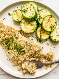 A baked haddock fillet next to zucchini on a white plate.