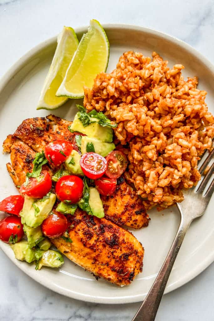 An overhead shot of a plate with tilapia, rice, limes, and a fork.