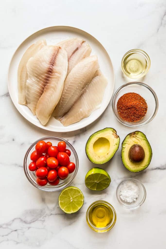 Tilapia fillets, blackening seasoning, an avocado, cherry tomatoes, lime, sea salt, and oil on a marble background.