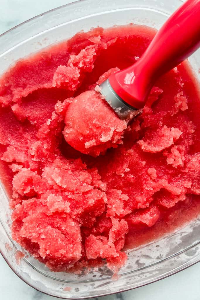 A closeup shot of watermelon sorbet in a large plastic container with an ice cream scoop.