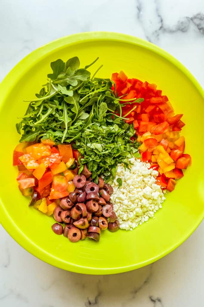 Arugula, chopped bell pepper, chopped olive, chopped tomatoes, and feta in a large green mixing bowl.