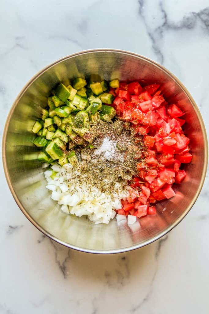 Chopped tomatoes, cucumbers, and white onion in a metal mixing bowl, topped with dried mint, sumac, pepper, and salt.
