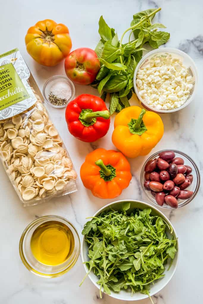 Bell peppers, tomatoes, a bag of pasta, olives, feta, arugula, fresh herbs, and olive oil on a marble background.