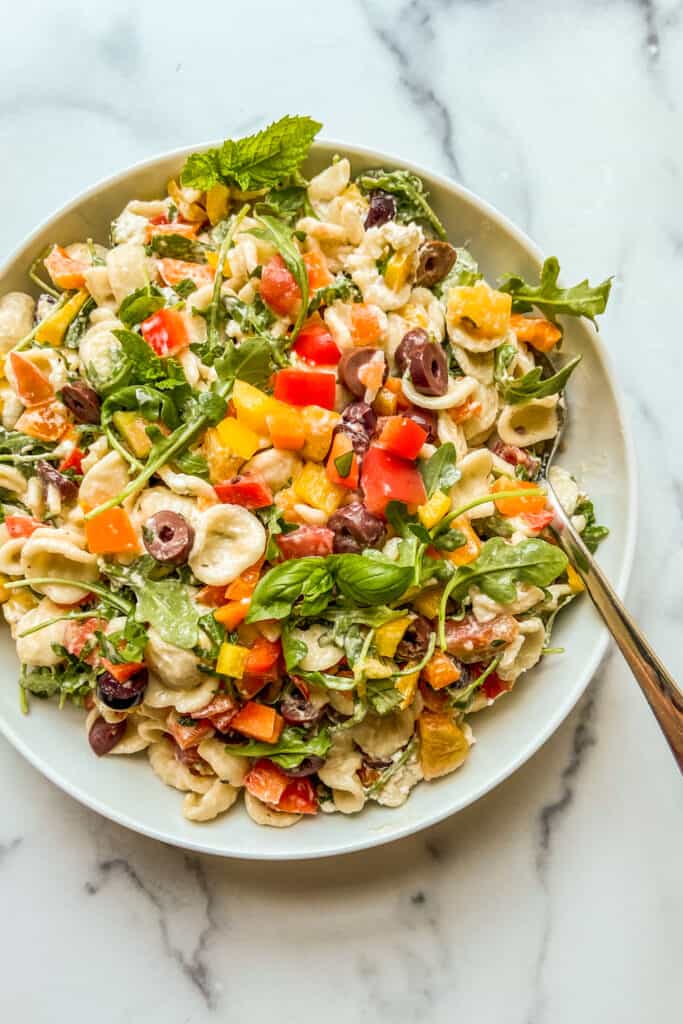 A pasta salad with bell peppers, olives, and arugula in a large white serving bowl.