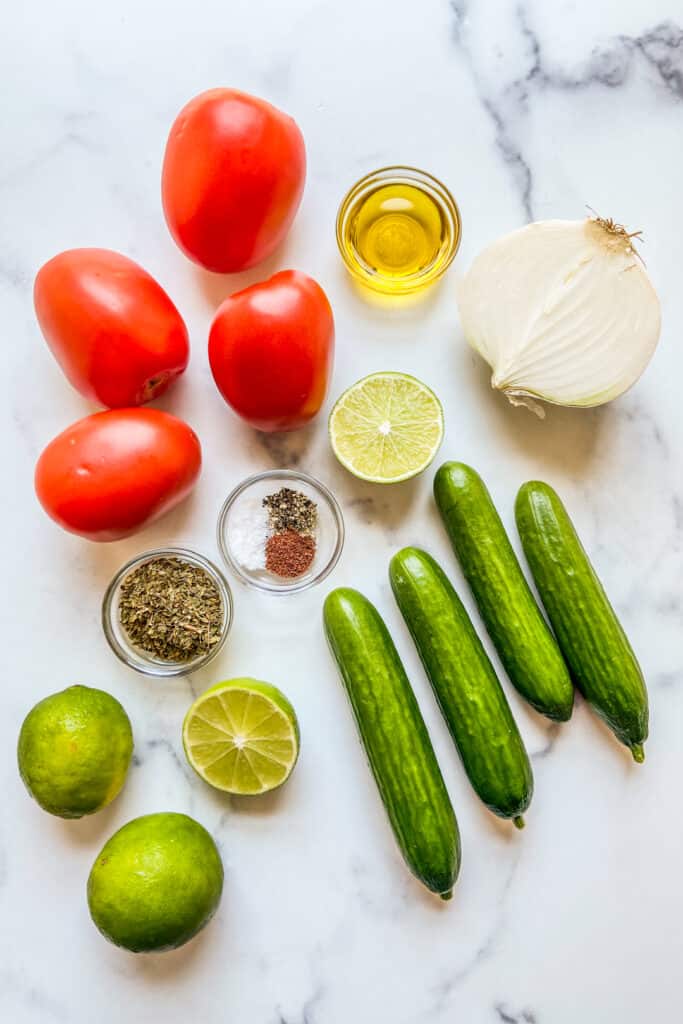 Persian cucumbers, Roma tomatoes, half a white onion, several limes, olive oil, dried mint, sumac, salt, and pepper on a marble background.