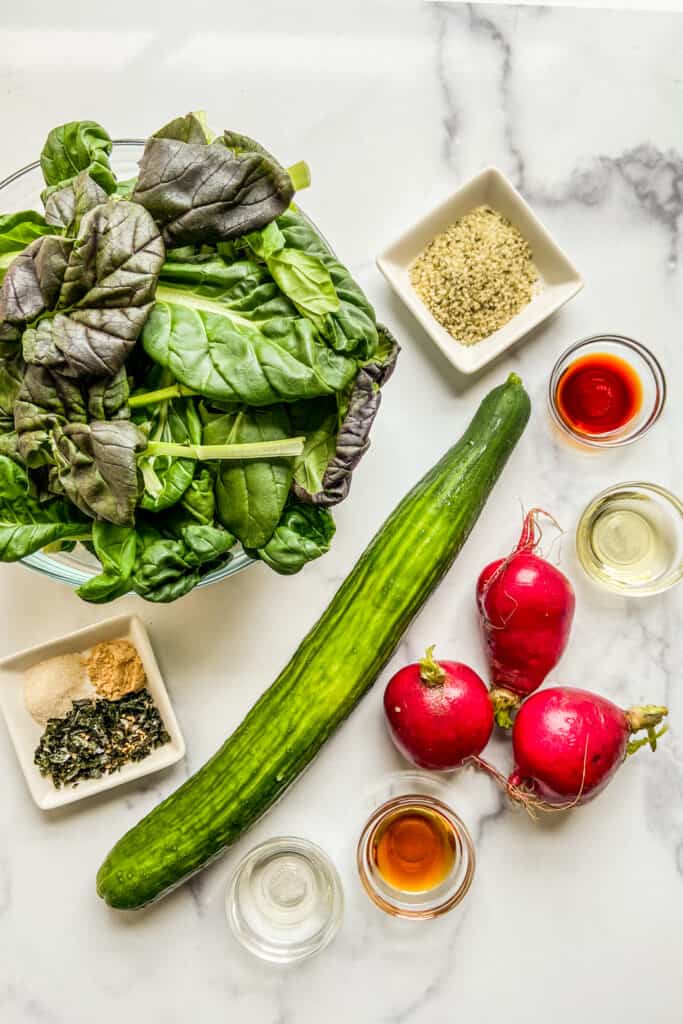 A bowl of tatsoi greens, an English cucumber, three red radishes, and bowls of hemps hearts, oils, furikake, ginger, soy sauce, and rice wine vinegar.