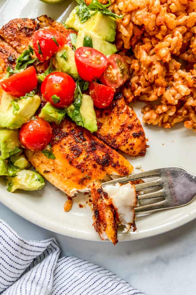 Blackened tilapia on a plate, topped with tomatoes and avocado, next to rice.