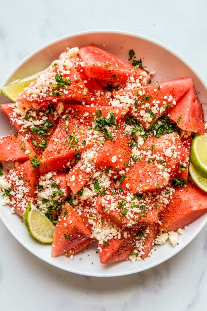 Sliced watermelon topped with cotija, tajin, and cilantro in a large white serving bowl.