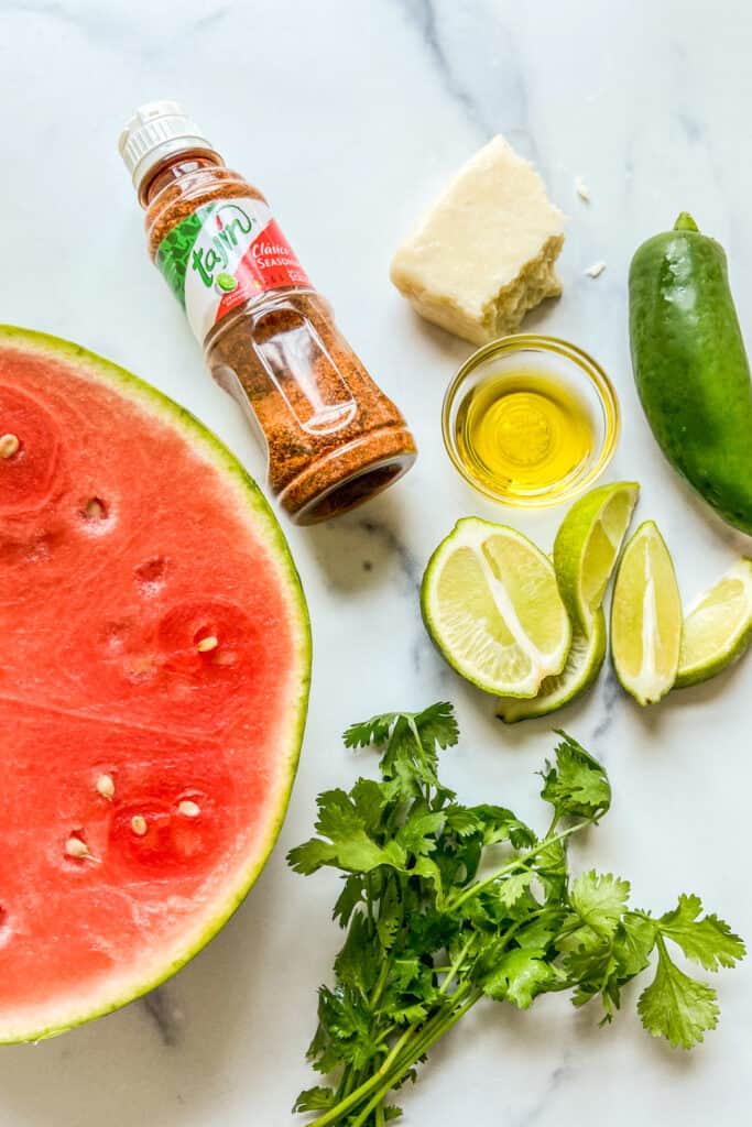 Half a watermelon, tajin seasoning, cut up limes, chunk of cotija cheese, a jalapeno, a small bowl of olive oil, and a small bunch of cilantro.