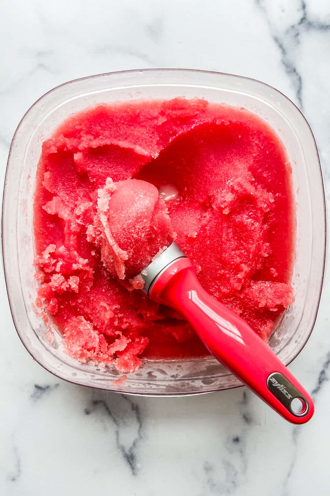 Watermelon sorbet in a large plastic container with an ice cream scoop.