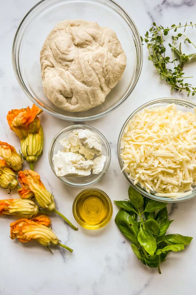 Pizza dough, shredded mozzarella, goat cheese, fresh herbs, zucchini flowers, and olive oil on a marble background.