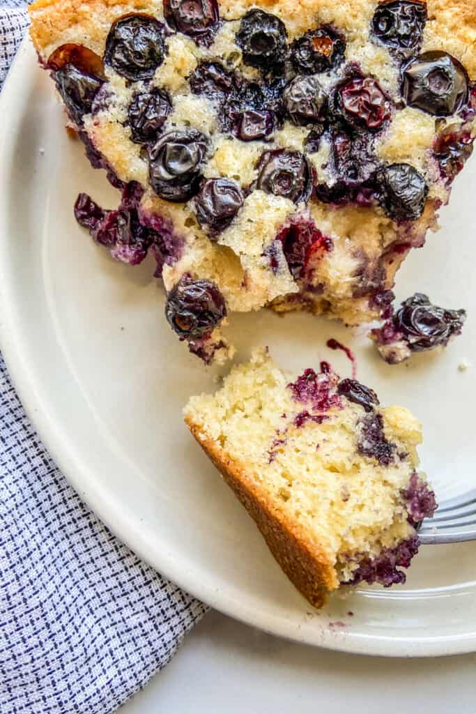 A closeup shot of a slice of blueberry cake with a piece cut off on a fork.