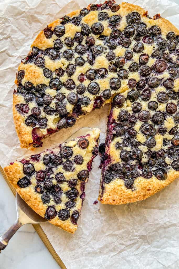 An overhead shot of a single layer yellow cake, filled with dozens of cooked blueberries. A triangle of cake is cut out and resting on a pie server. 