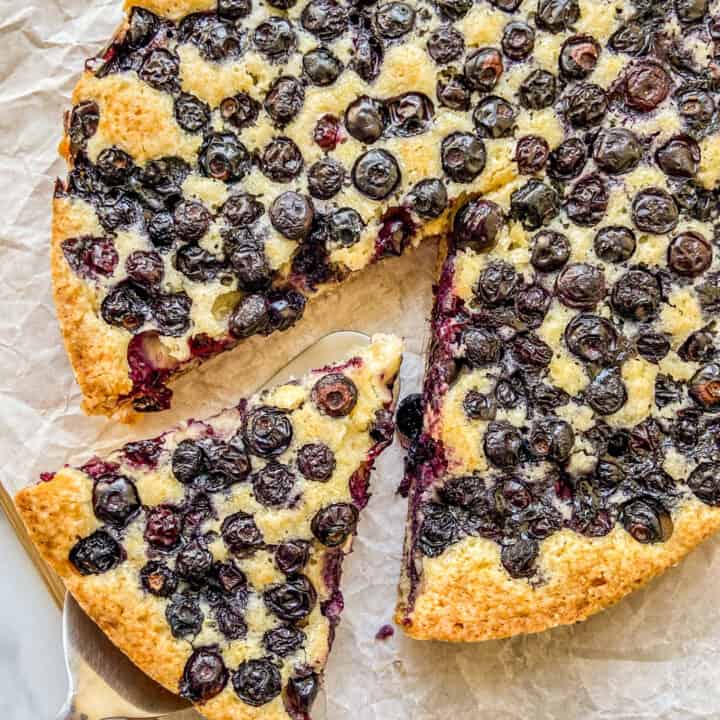 An overhead shot of a single layer yellow cake, filled with dozens of cooked blueberries. A triangle of cake is cut out and resting on a pie server.