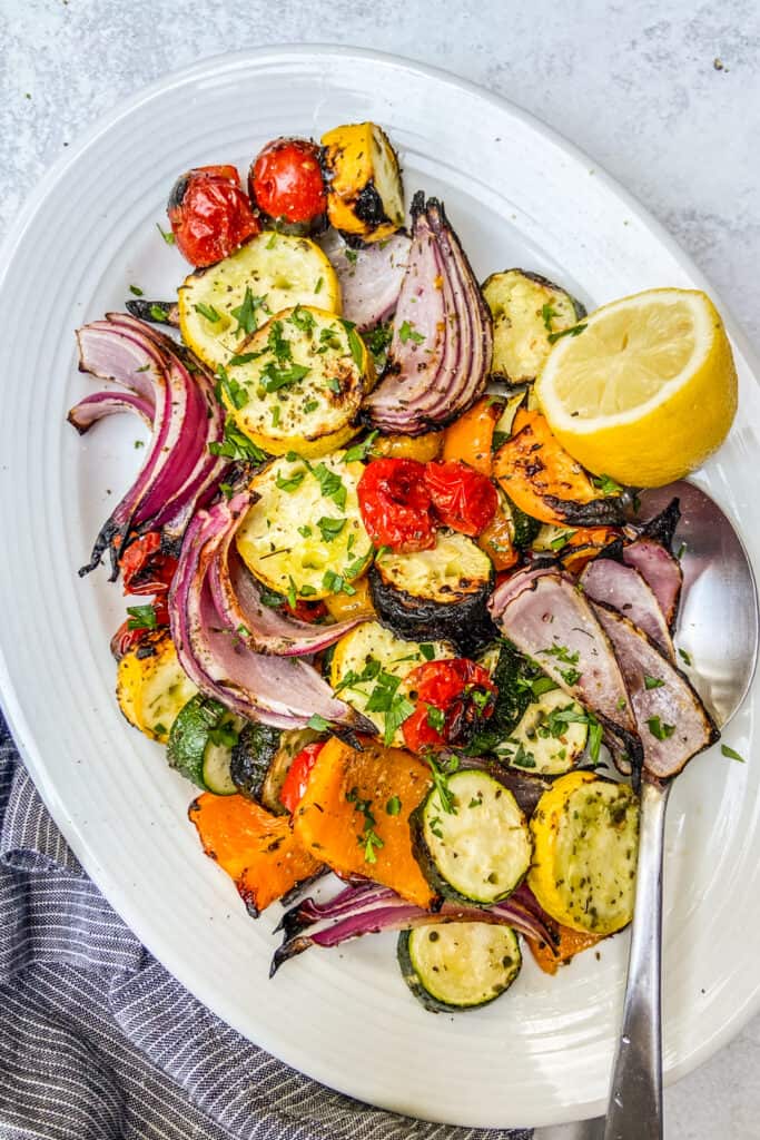 An overhead shot of a platter of grilled vegetables, topped with finely chopped herbs, next to a blue napkin.