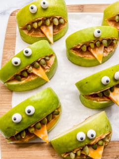 Apples slices with candy eyes, cheese tongues, and pumpkin seed teeth on a cutting board.