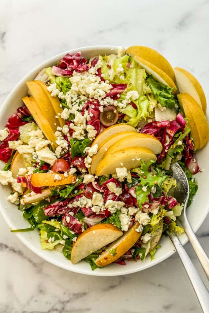 An overhead shot of an Asian pear salad in a white bowl with silver serving spoons.