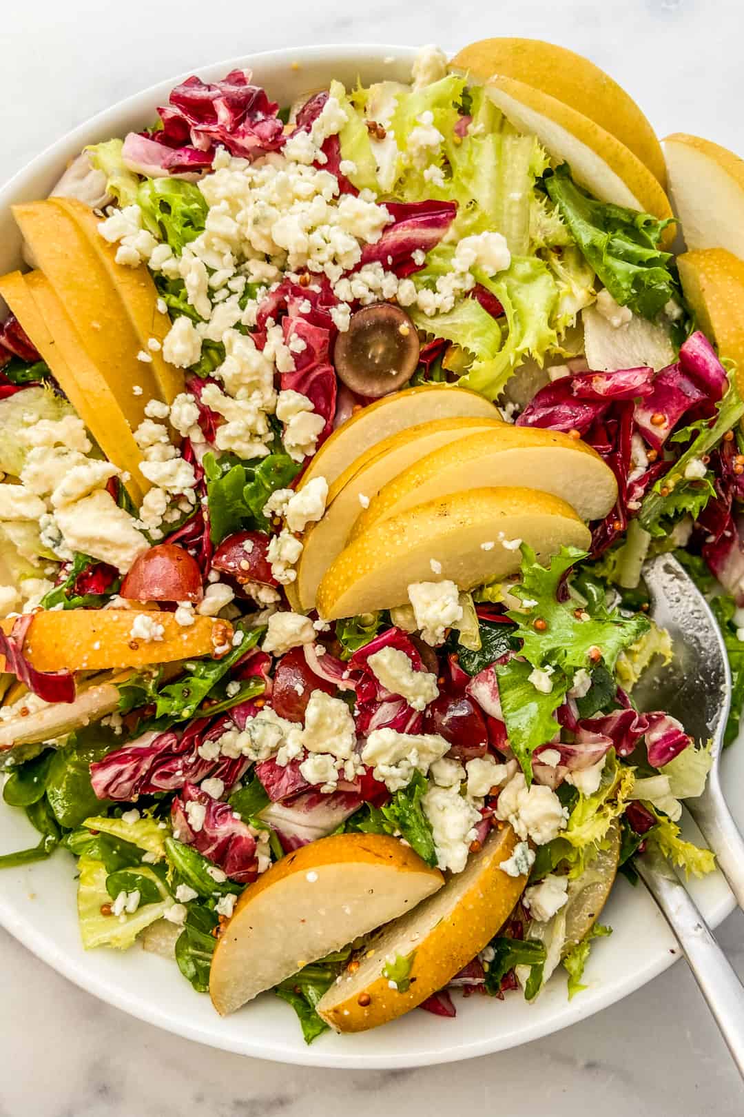 A closeup shot of an Asian pear salad with arugula, blue cheese, and grapes in a white serving bowl.