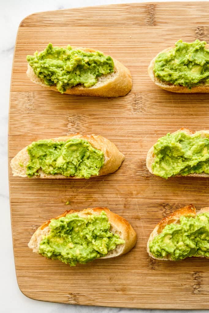 Avocado spread on sliced of French bread, sitting on a cutting board.