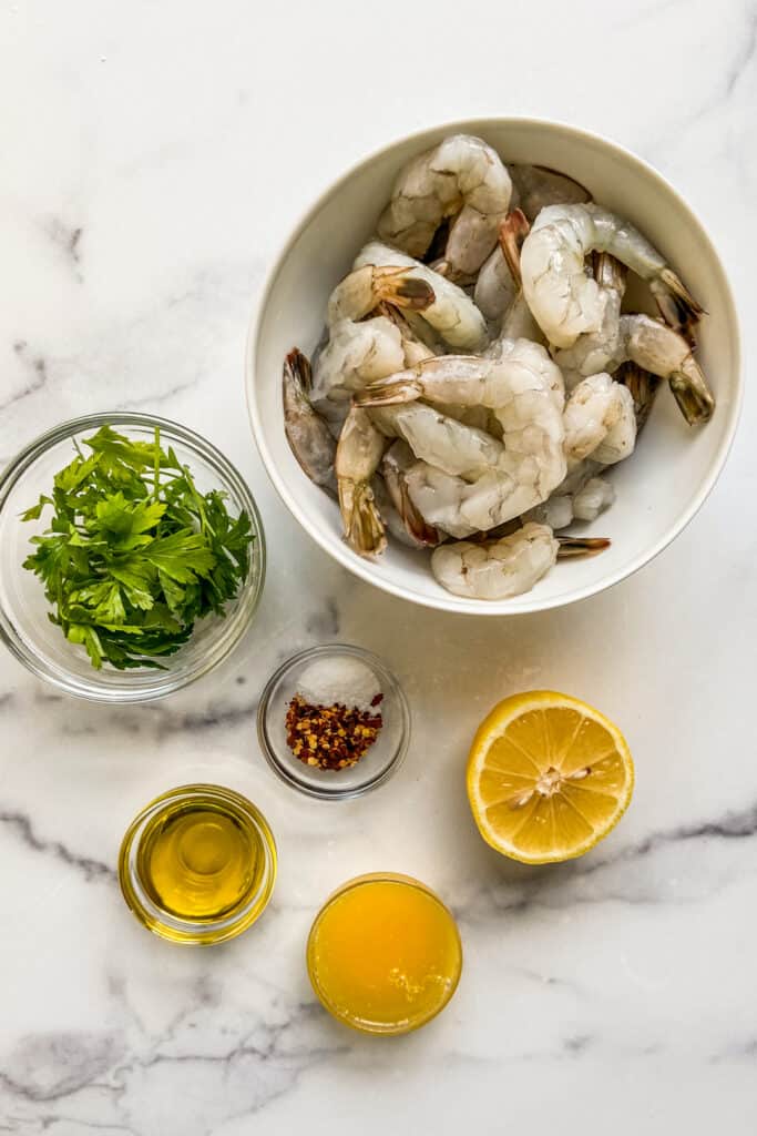 Shrimp, parsley, red pepper flakes, olive oil, and a sliced lemon on a marble backdrop.