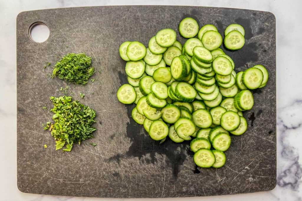 Sliced cucumbers and chopped dill and mint on a black cutting board.