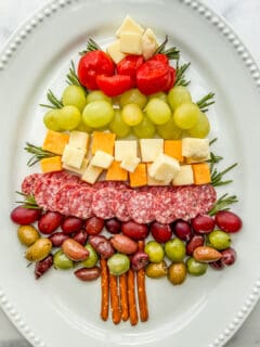 An overhead shot of a Christmas tree made out of cheese, fruit, and salami on a large white platter.
