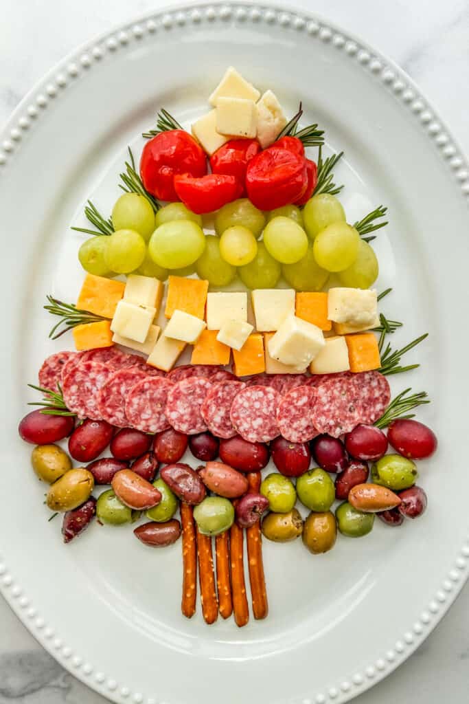 An overhead shot of a Christmas tree made out of cheese, fruit, and salami on a large white platter.