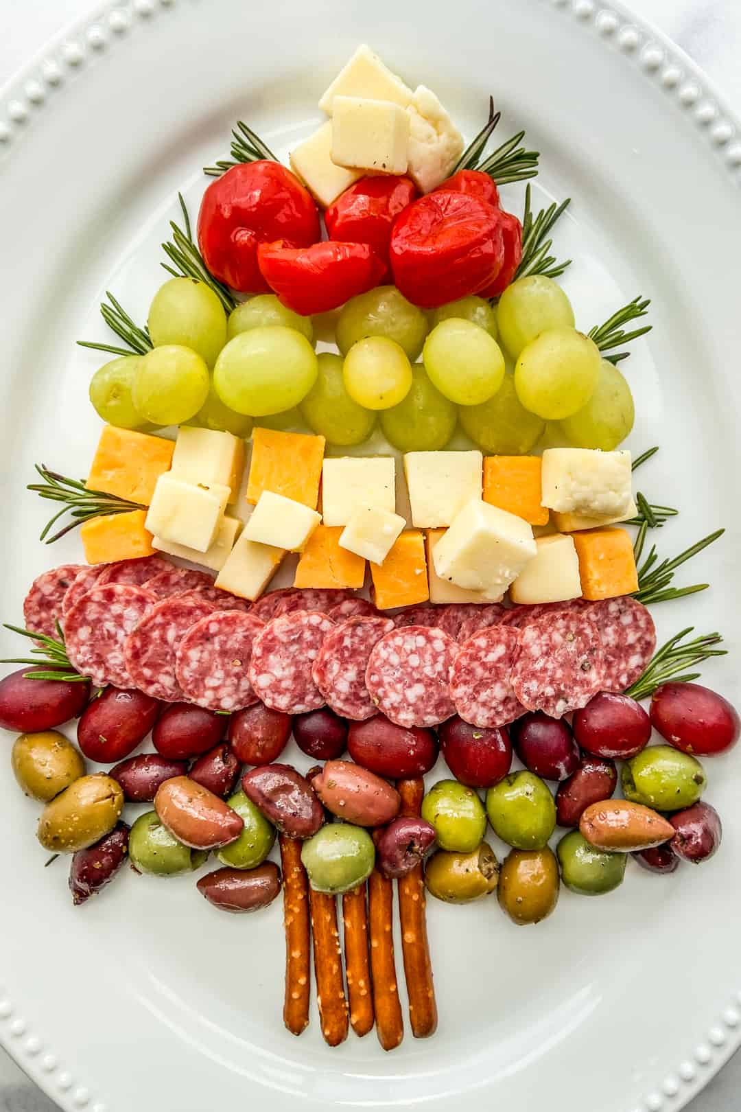 An overhead shot of a Christmas tree made out of cheese, fruit, and salami on a large white platter.