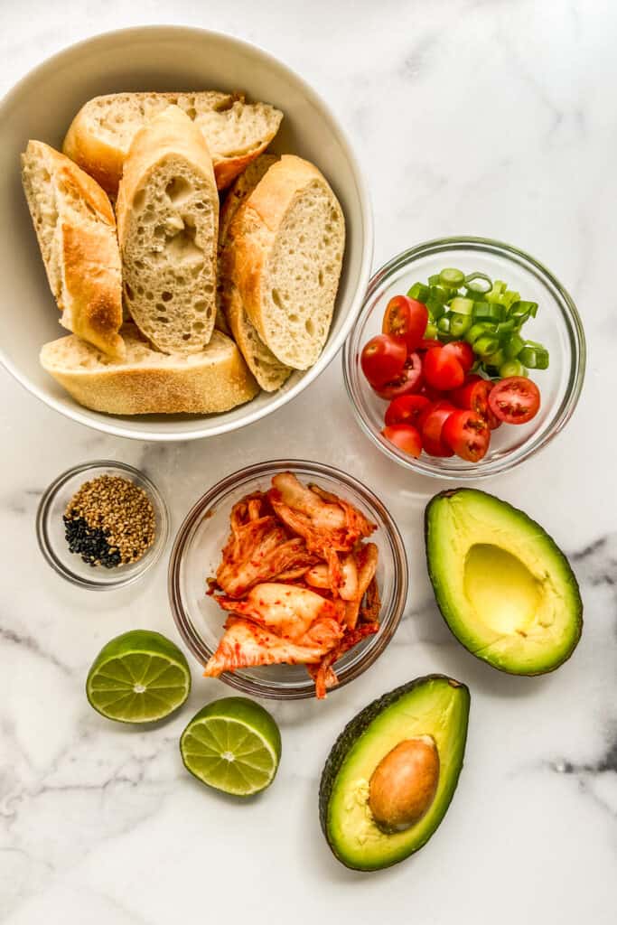 Sliced French bread, avocado, kimchi, cherry tomatoes, diced green onions, lime, and sesame seeds on a marble background.