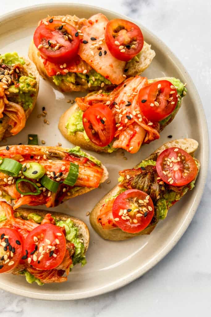 An overhead shot of a plate of crostinis topped with avocado, kimchi, cherry tomatoes, green onions, and sesame seeds.