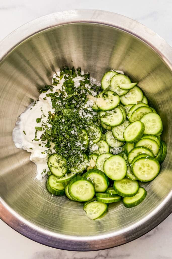 Sliced cucumbers, Greek yogurt, herbs, and sea salt in a large, metal mixing bowl.